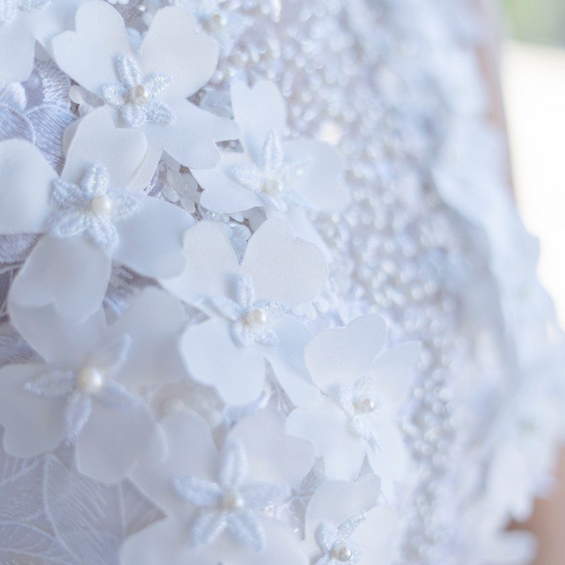 Close-up of hand-sewn pearls and floral appliqués on lace fabric of wedding dress, showcasing artisanal craftsmanship.
